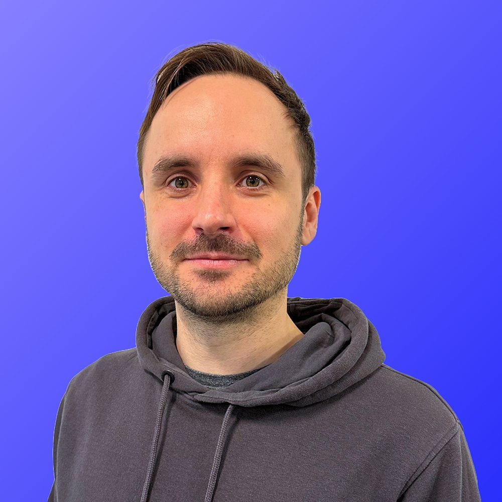 A man wearing a black sweater and a blue shirt is standing in front of a blue background.