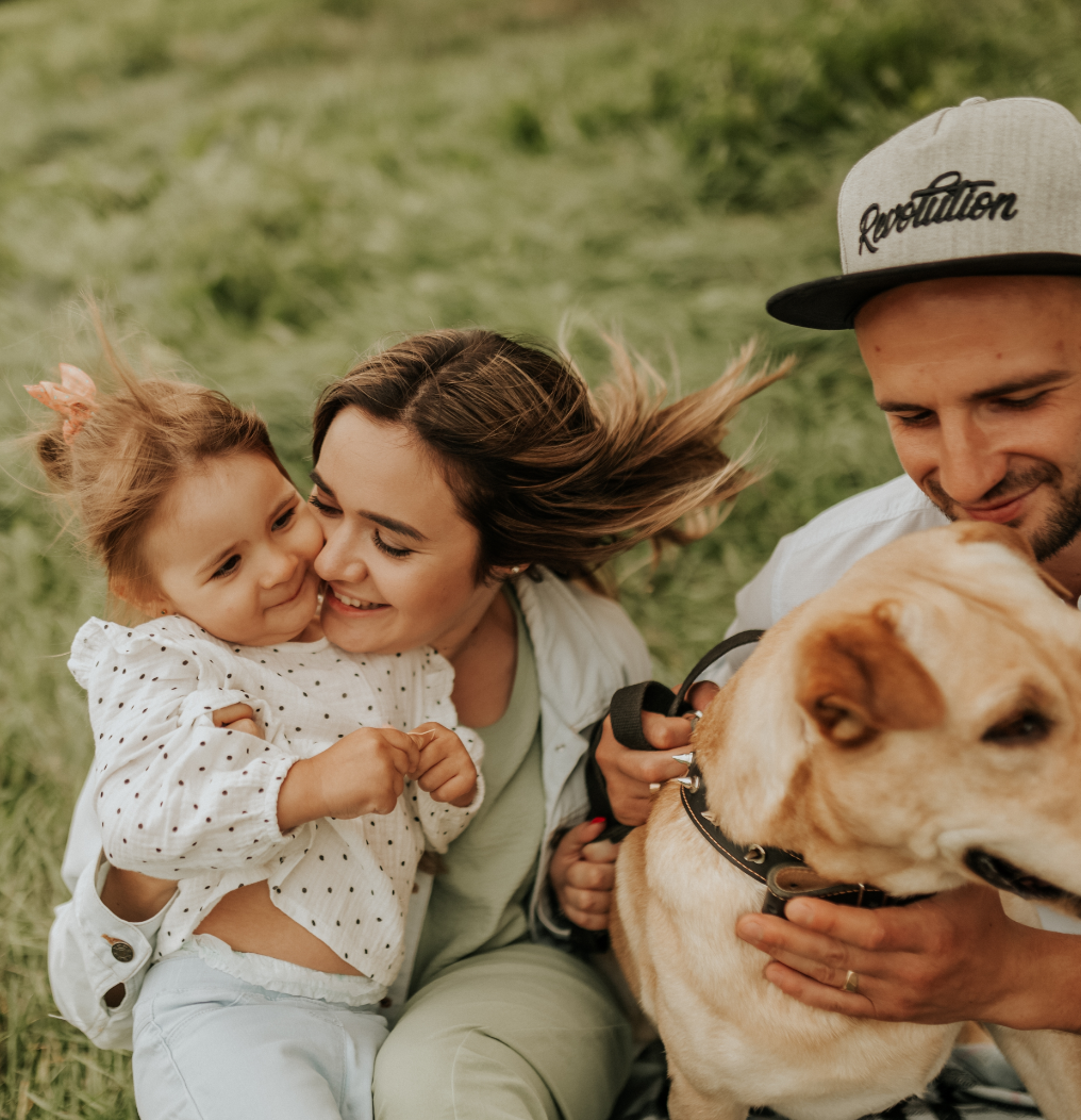Family sitting on grass, cuddling a toddler and a large dog outdoors.