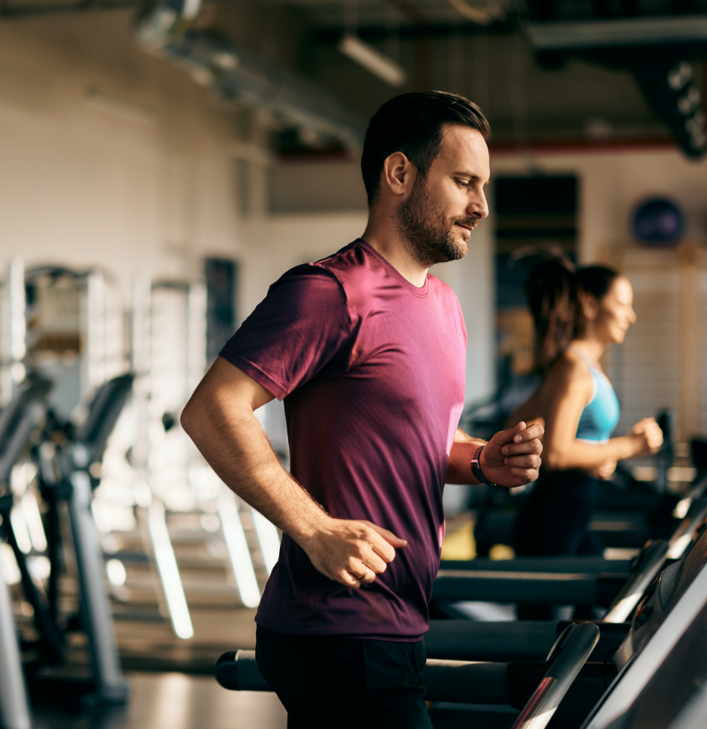 Man running on a treadmill in a gym, wearing a purple shirt with blurred exercisers behind him