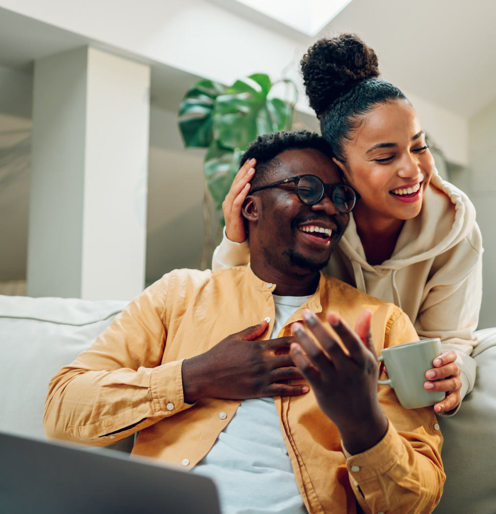 Two people smiling on a couch, sharing a mug in a bright living room