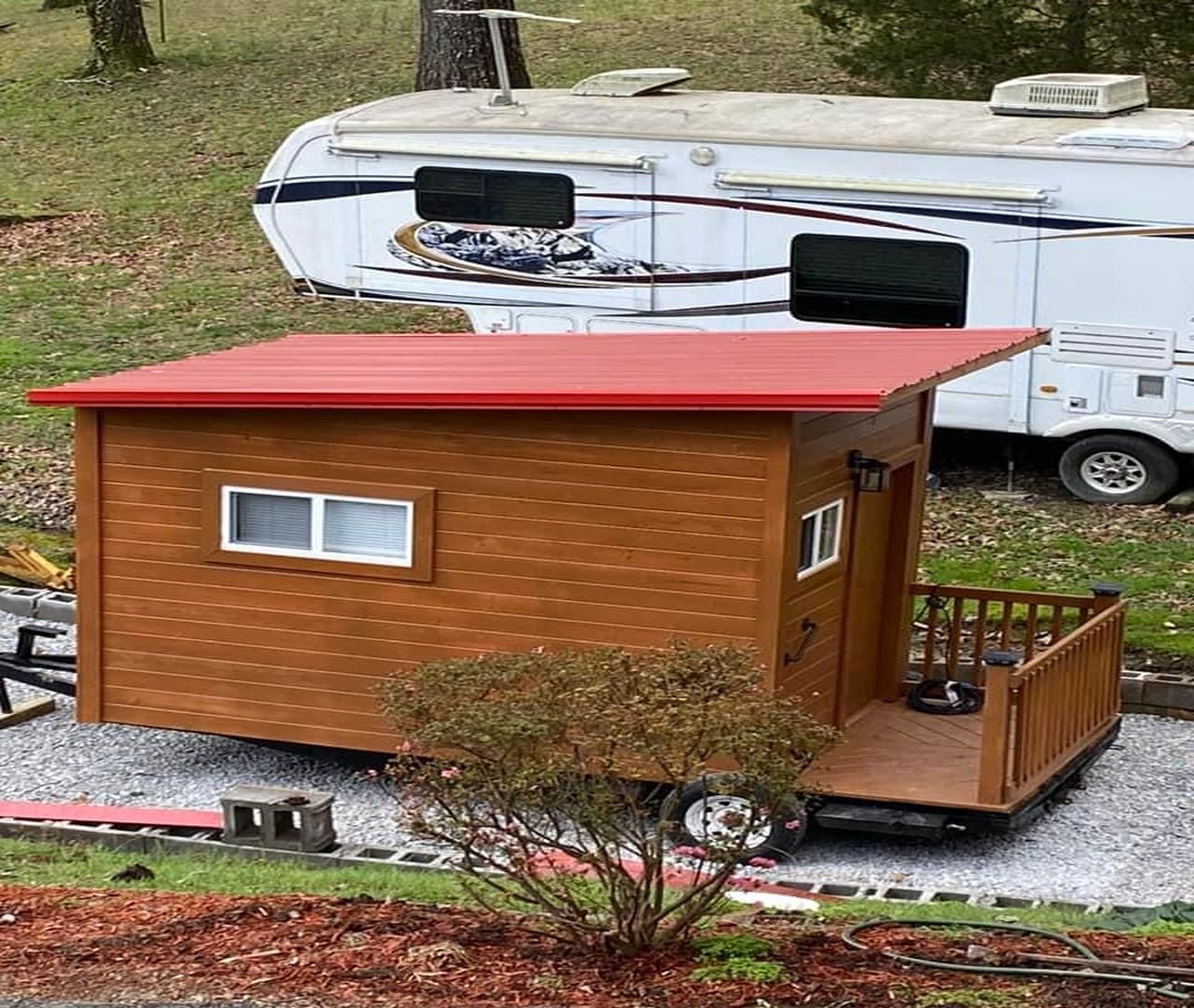 Small brown tiny house with a red roof beside a white RV in a grassy area