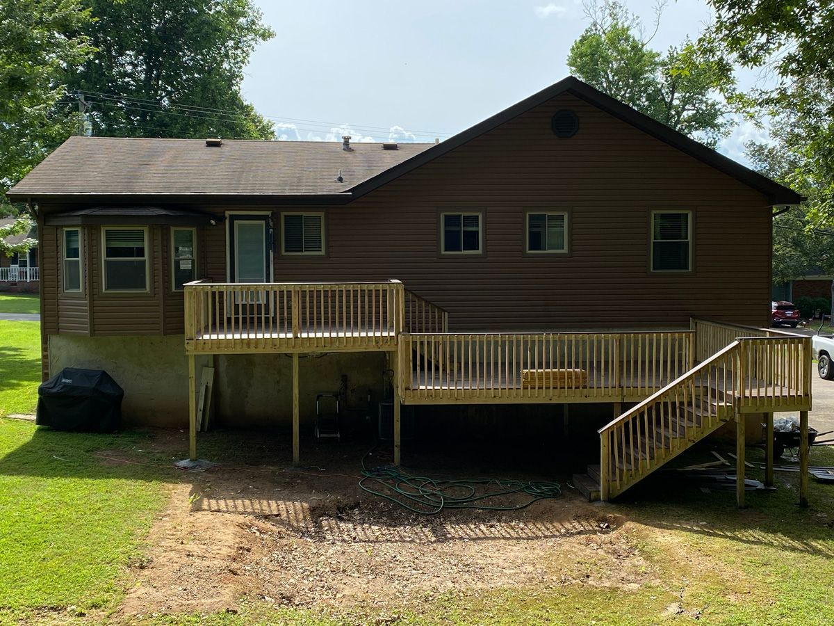 Wooden cabin with a front deck and stairs in a grassy yard, surrounded by trees.