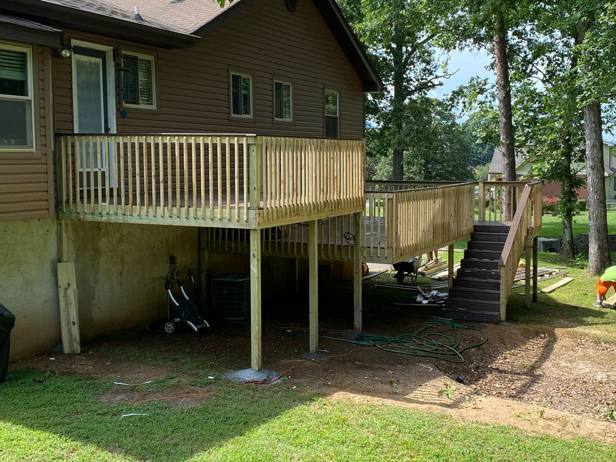 House with raised wooden deck and stairs, with a stroller stored underneath in a wooded yard.