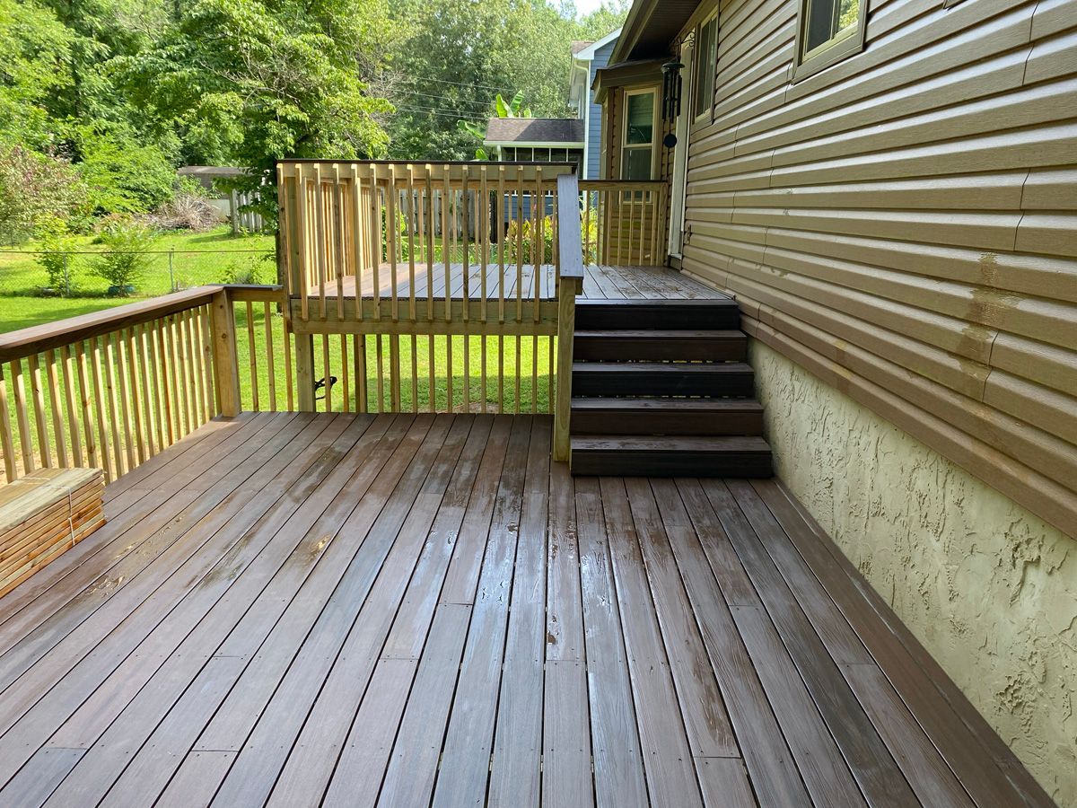 Wooden backyard deck beside a house, with railing, stairs, and green trees beyond.