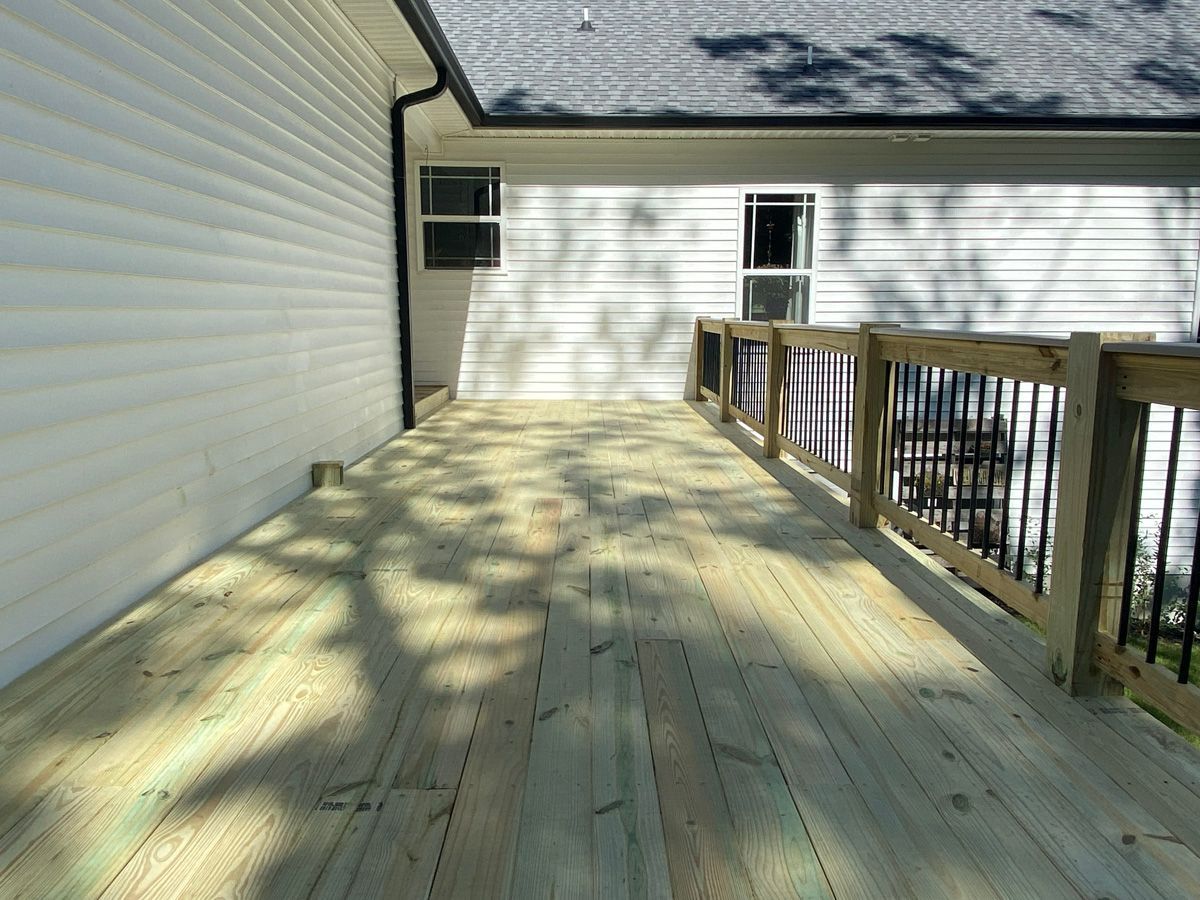 Wooden deck walkway beside a white house, with railing and dappled sunlight.