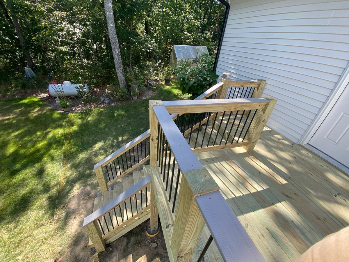 Wooden backyard deck with stairs and railing beside a white house, overlooking a grassy yard.