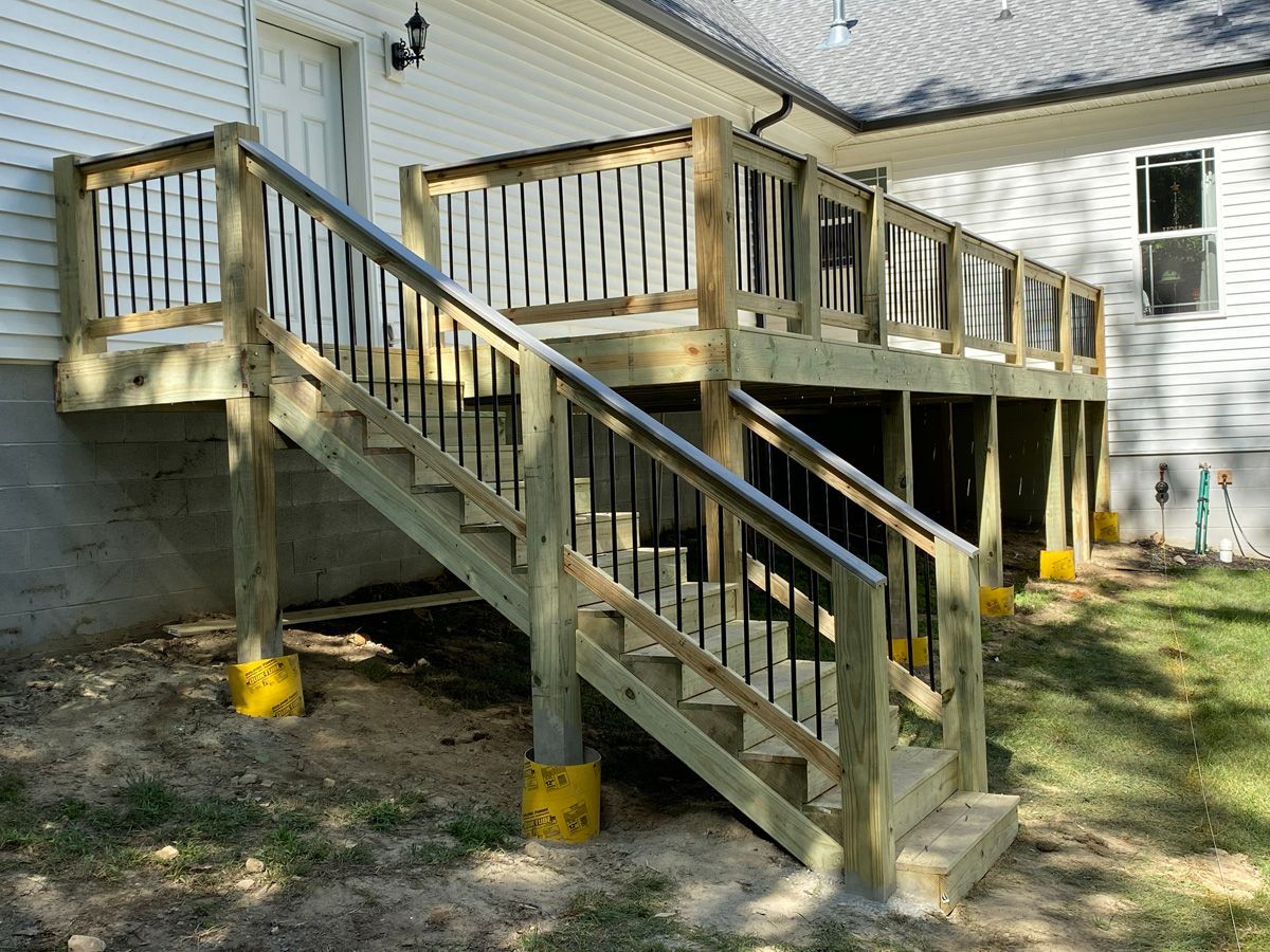 Raised wooden deck with stairs and railings beside a house, supported by concrete posts with yellow covers