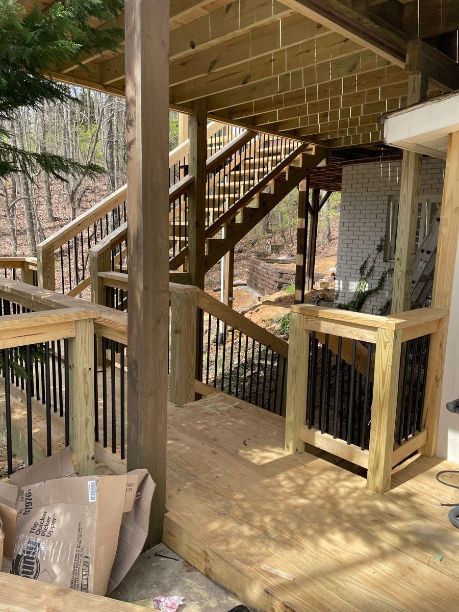 Wooden deck with stairs and railing beneath a house, surrounded by trees.