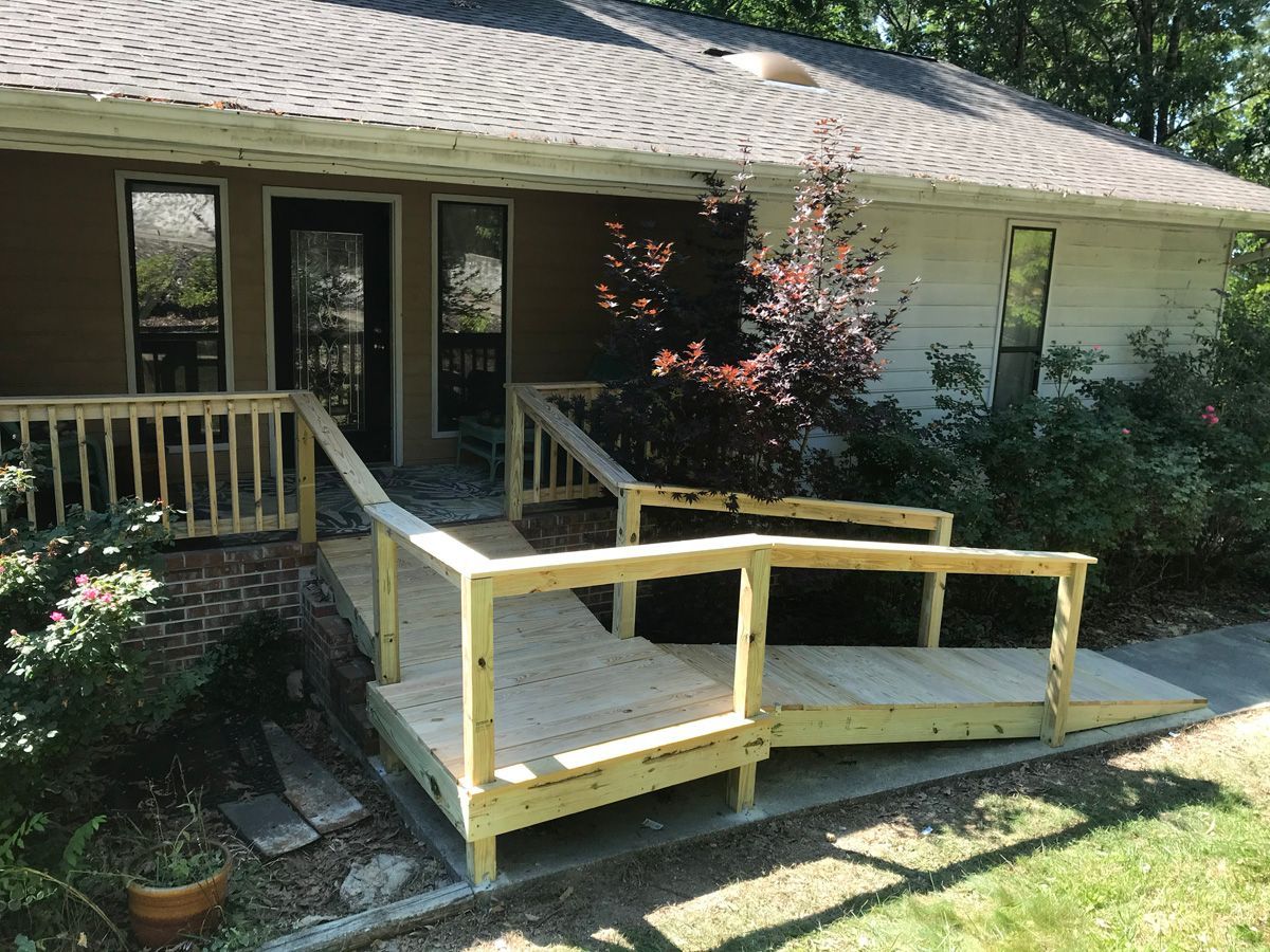 Wooden wheelchair ramp leading to a house porch with dark doors and windows, shaded by trees.