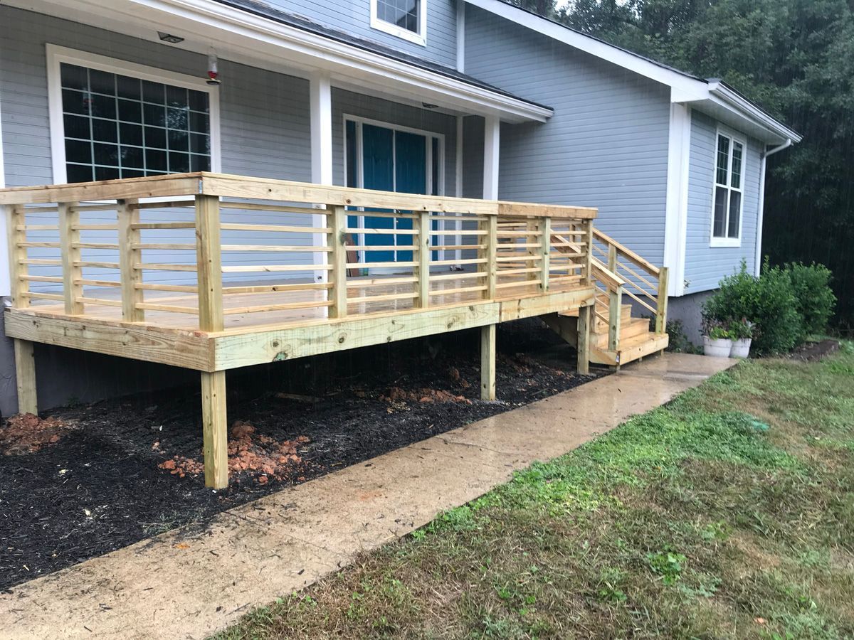 Wooden front porch ramp with railings attached to a gray house, beside a gravel path and grass.