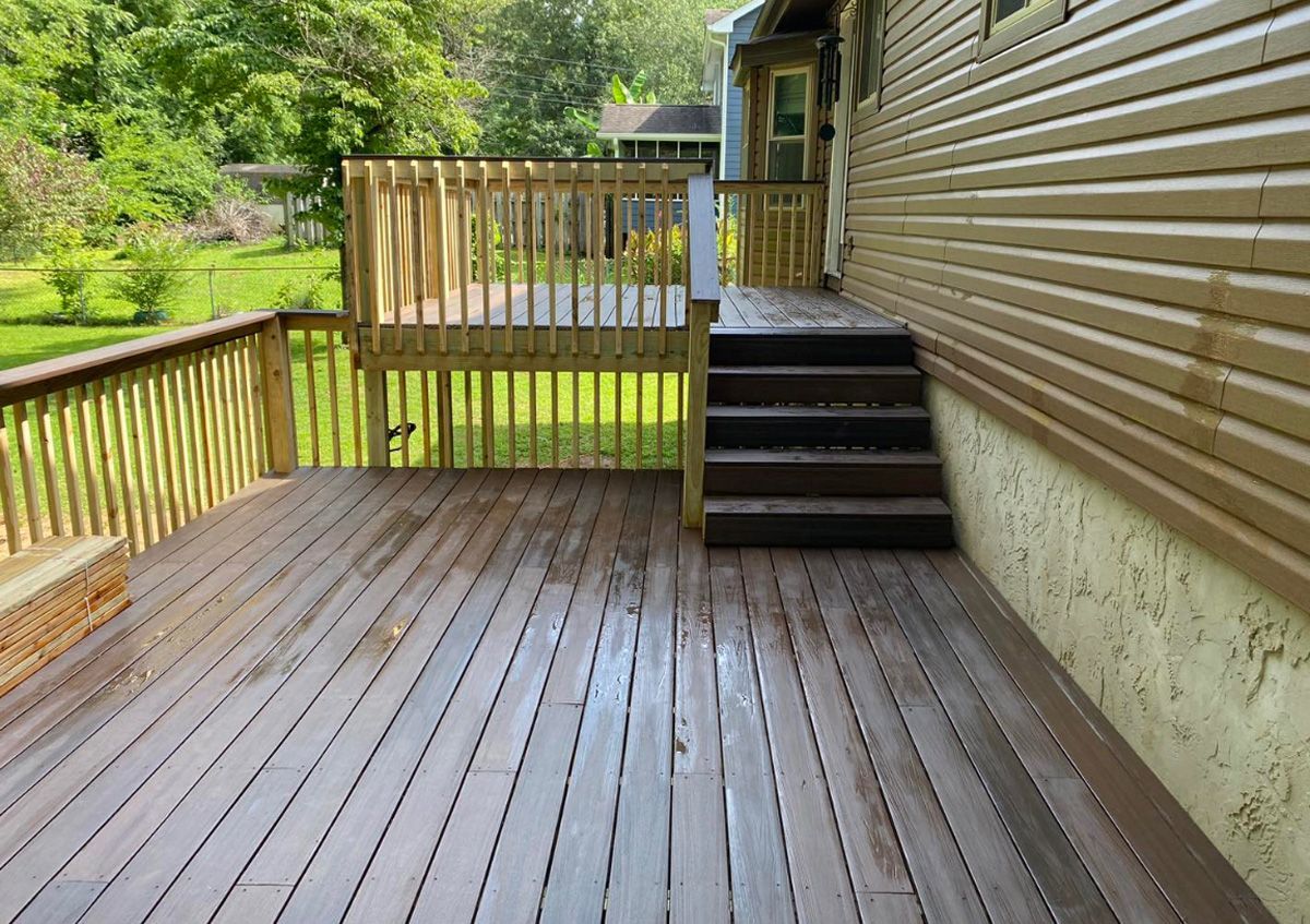 Wooden backyard deck with railing, steps, and a house exterior beside a green yard and trees.