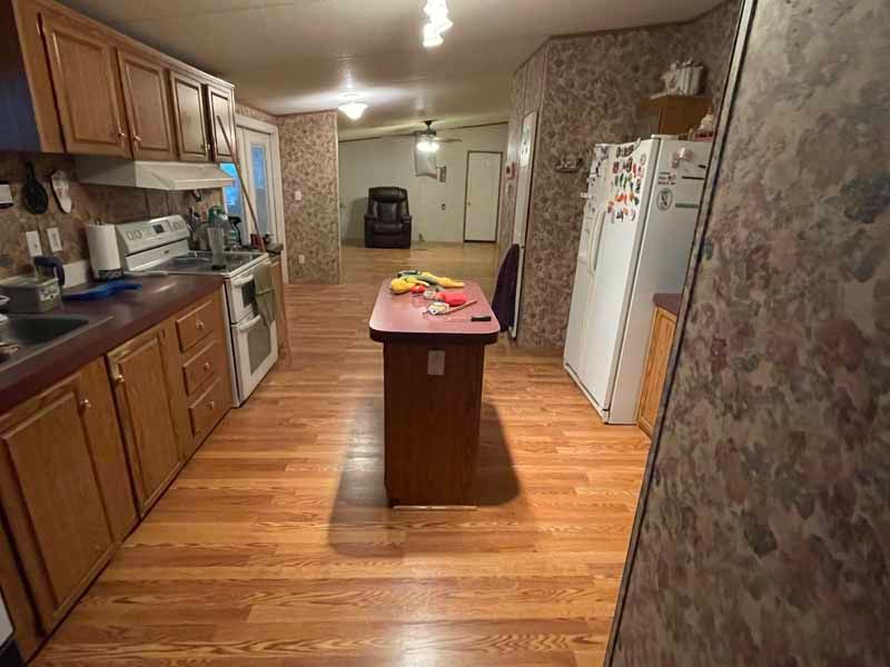 Empty kitchen with wood floors, central island, wooden cabinets, and white appliances, viewed toward a doorway.