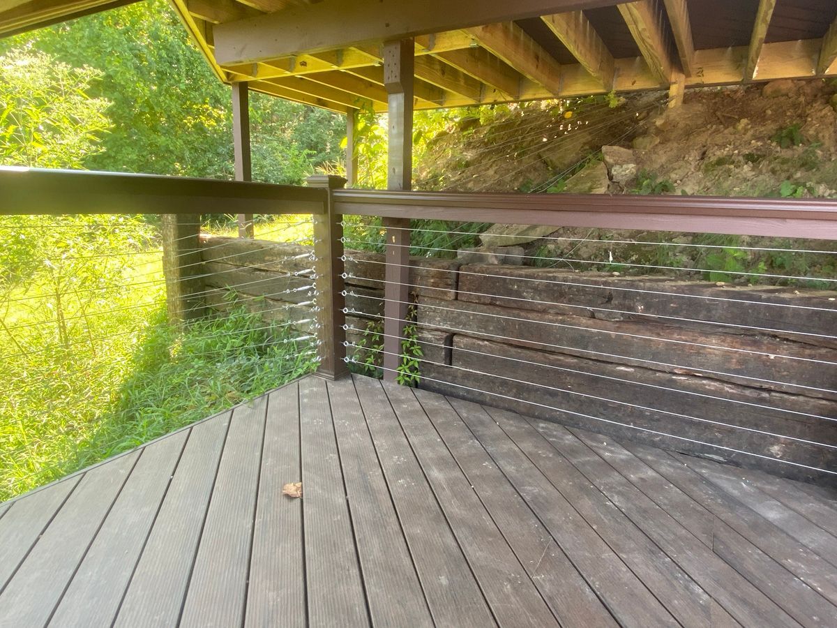 Wooden deck under a covered roof beside a grassy hillside, with black railing and sunlit greenery.