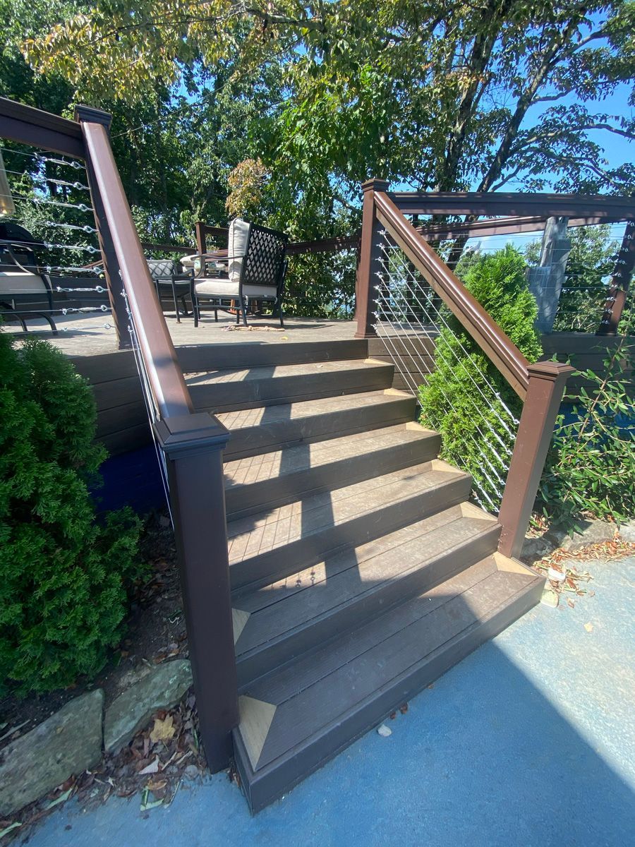 Outdoor wooden stairs with brown railings leading up to a deck beside greenery and a blue path