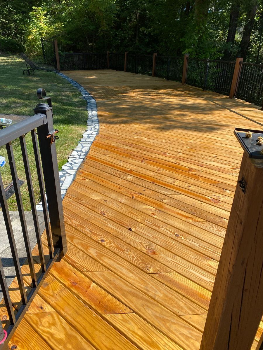 Wooden deck walkway with black railing, bordered by grass and trees in a sunny yard.