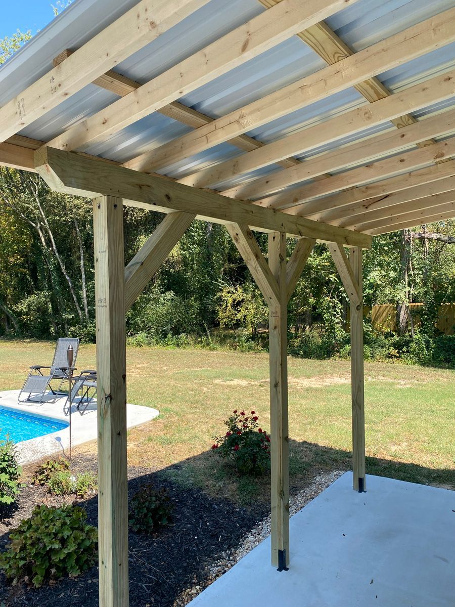 Wooden pergola with open rafters beside a backyard pool and patio, overlooking a grassy yard.