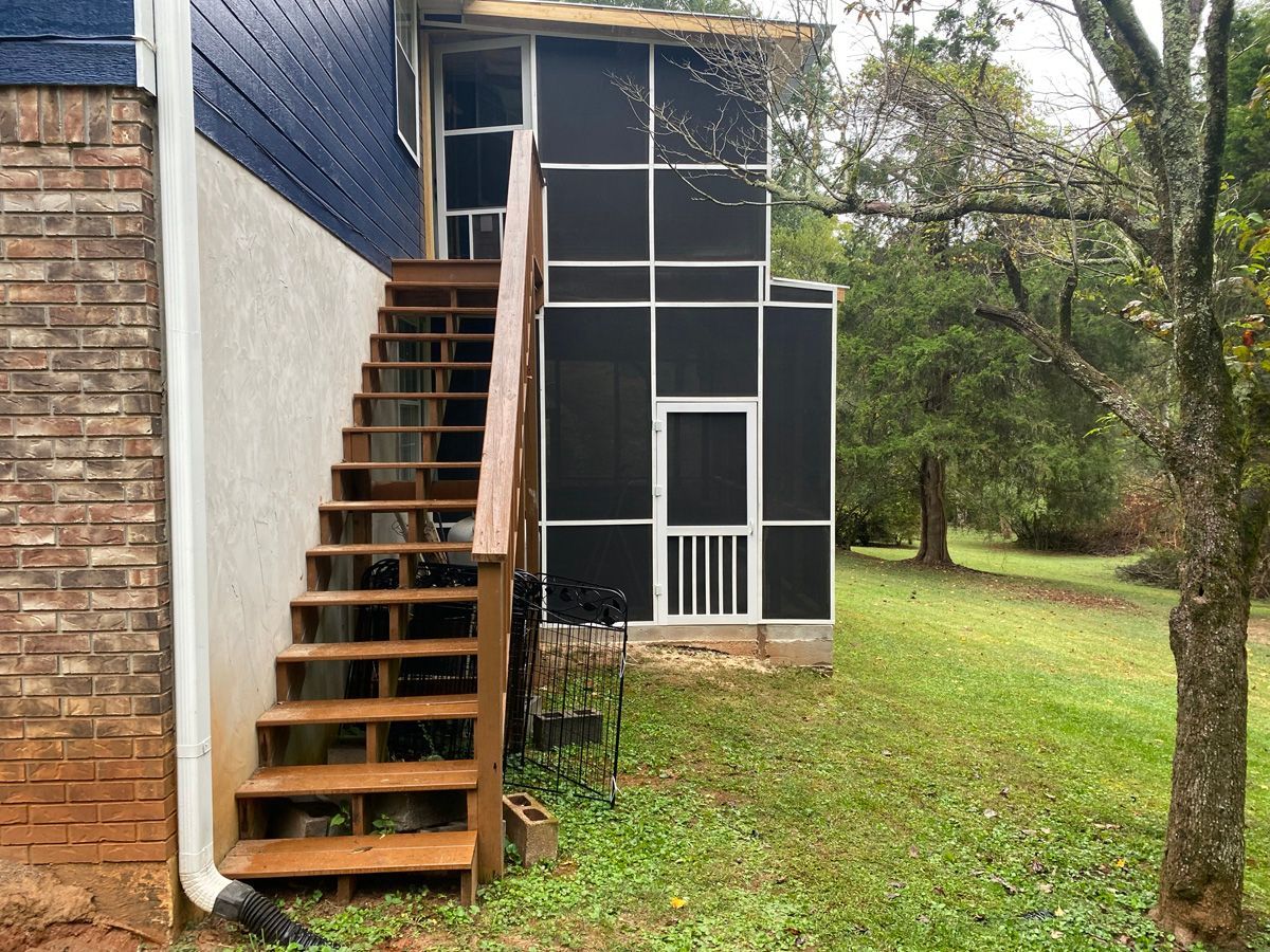 Wooden stairs on the side of a house leading to an upper door, beside a screened porch and grassy yard.