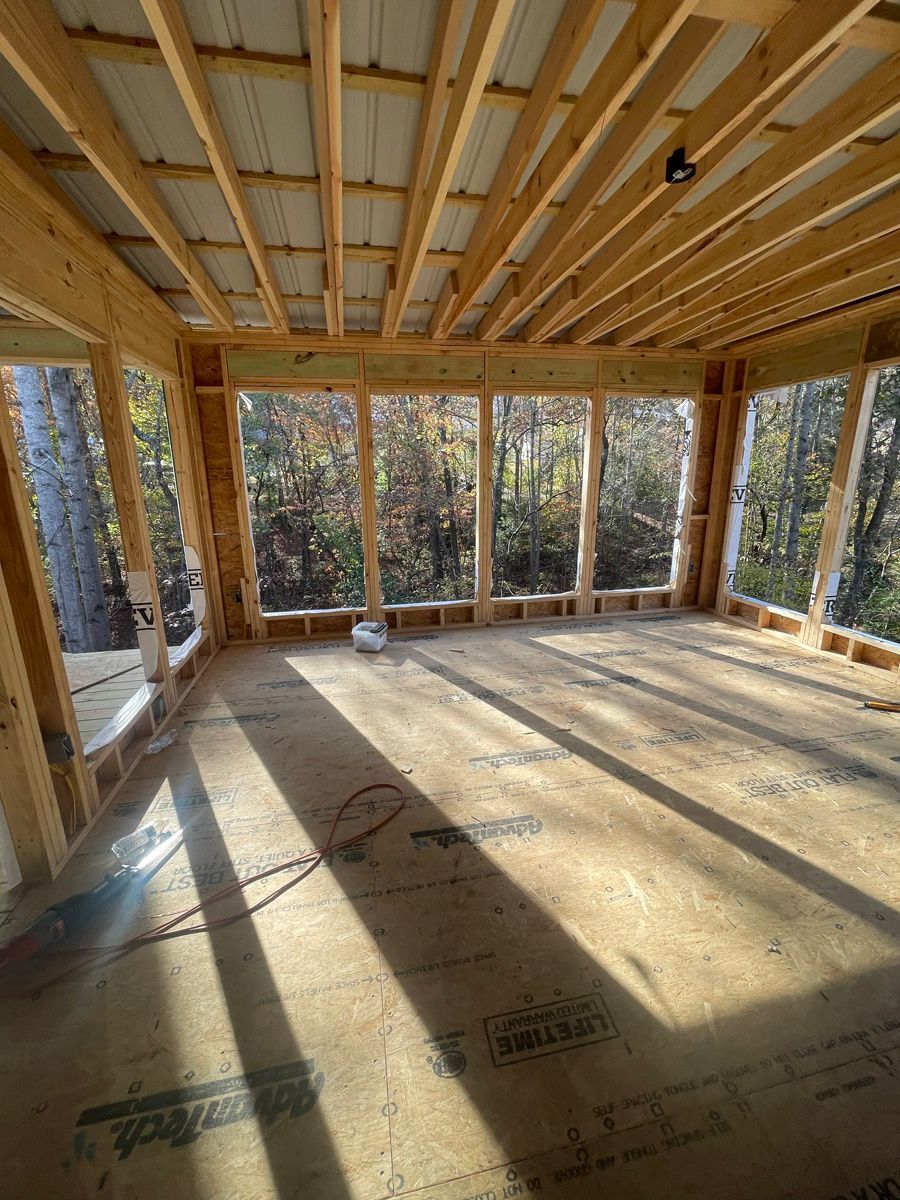 Framed house interior with exposed wooden studs, floor joists, and sunlight streaming through open wall bays