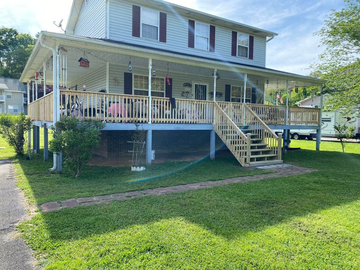 White two-story house with a wraparound porch and stairs, raised on stilts over a grassy yard.