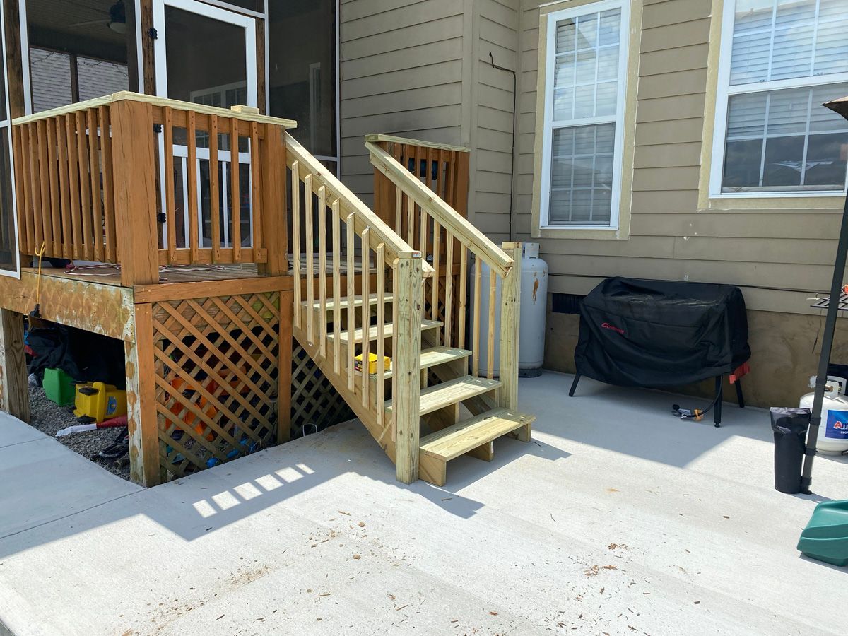 Backyard deck with wooden stairs and railing beside a house, with a grill on the patio.