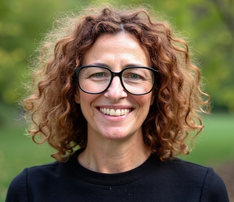 Smiling person with curly hair and glasses wearing a black top outdoors with a green blurred background — AT My Place Counselling and Psychotherapy in Neutral Bay, NSW
