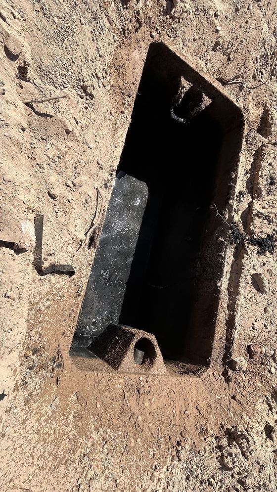 Close-up of a rusted rectangular metal opening in a dirt wall with a small pipe at the bottom.