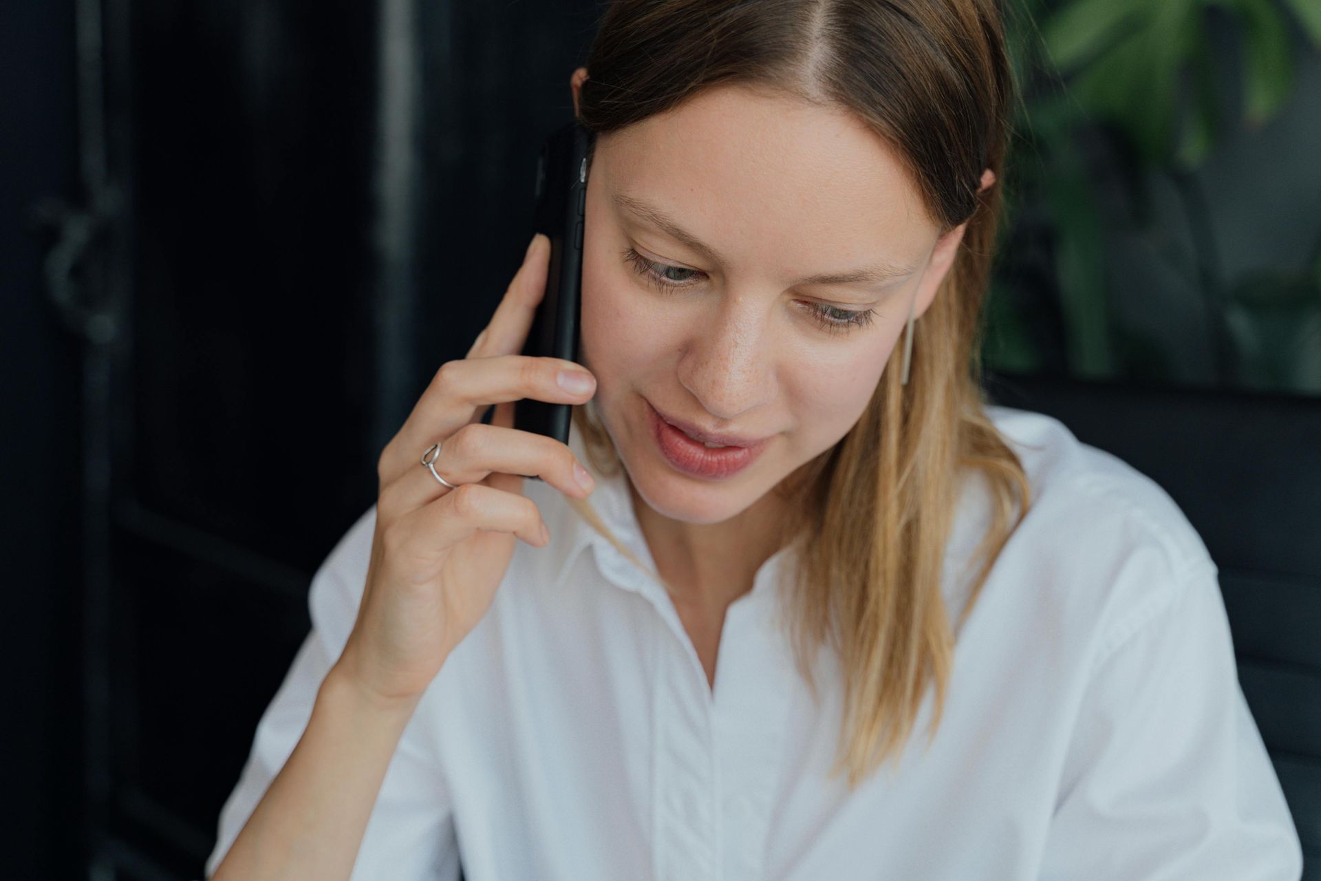 Woman in a white shirt talking on a phone, looking down with a slight smile indoors.