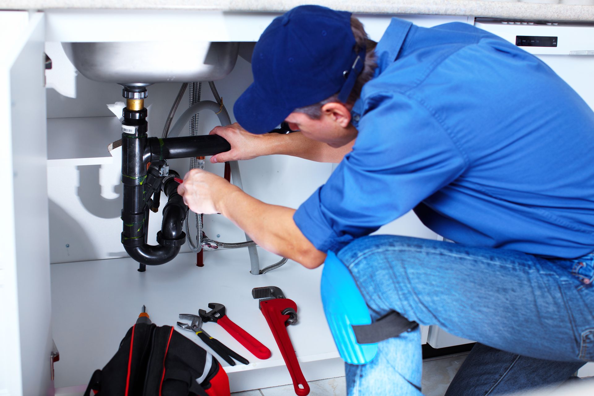 Plumber in blue uniform, kneeling under a sink, working with tools.