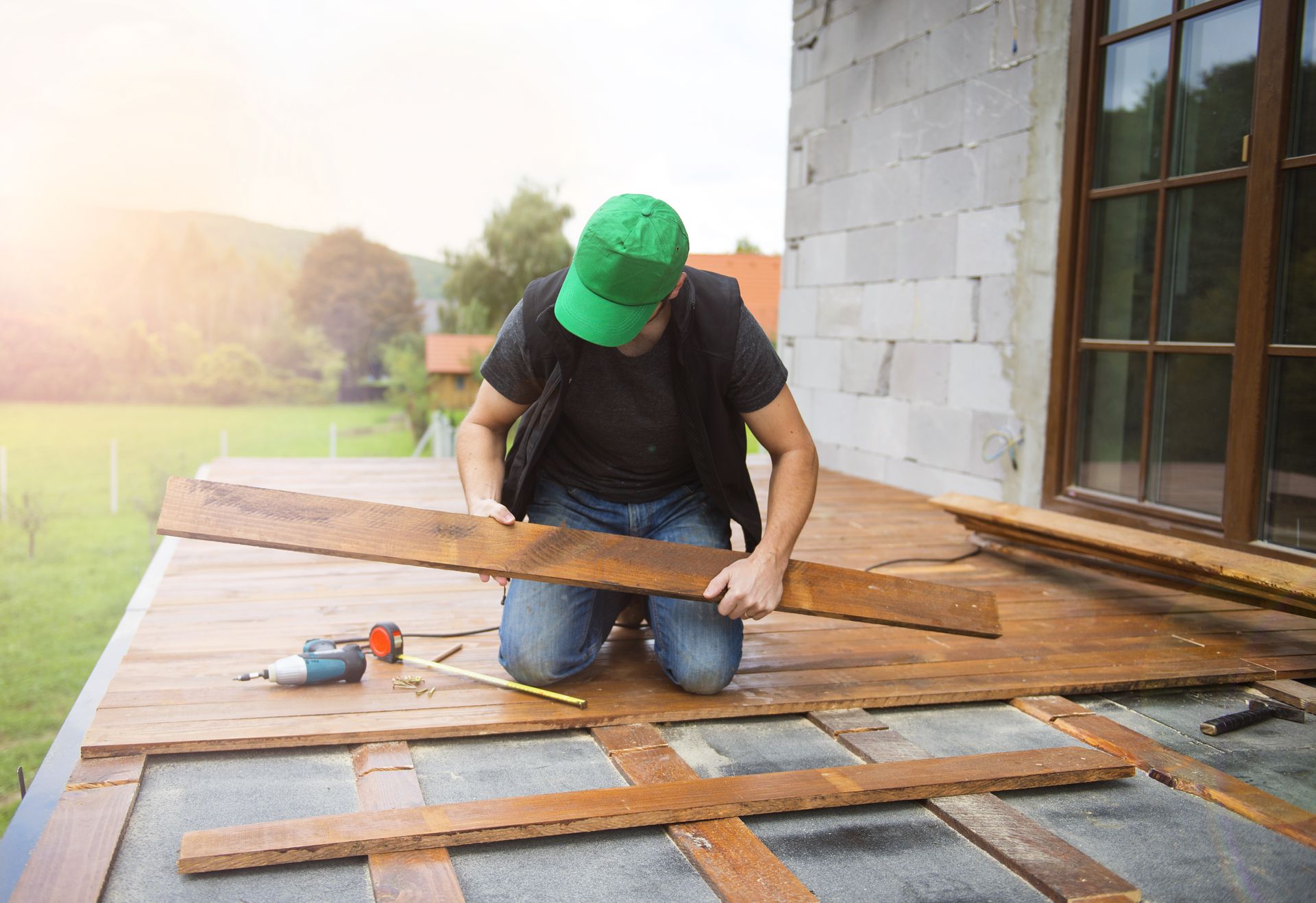 Person kneeling on deck, holding wood plank, installing it. A power drill and tools nearby. Green cap, sunny outdoors.
