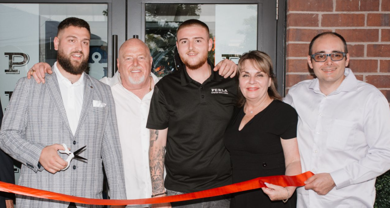 Five people posing at a ribbon-cutting event, holding a red ribbon indoors.