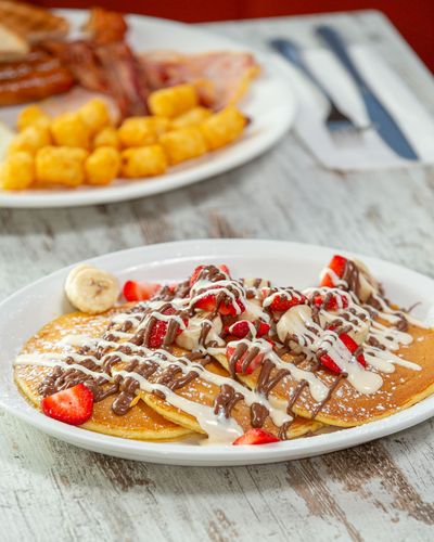 Chocolate-drizzled crepes with strawberries and banana on a white plate, with tater tots and breakfast in the background