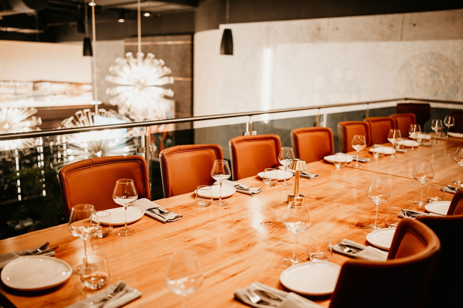 Long wooden dining table set with orange chairs, plates, glasses, and centerpieces in a modern restaurant