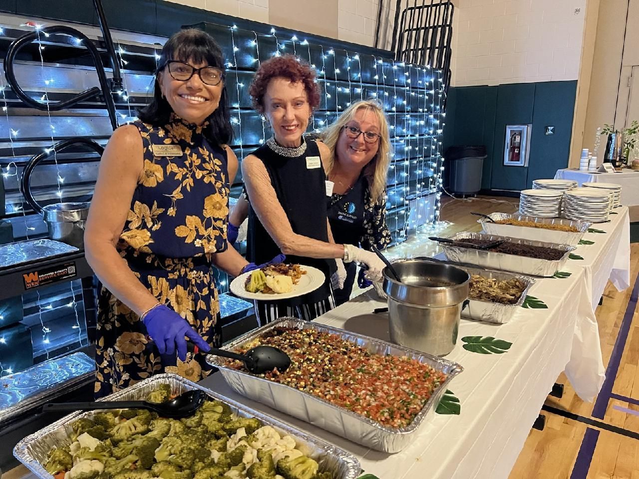 Three women at a buffet table. One serves food. Decorated hall with string lights.