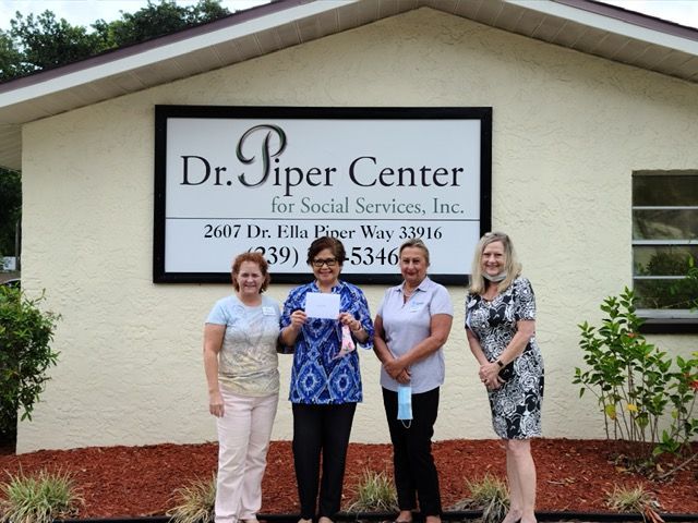 Four women stand outside the Dr. Piper Center, holding a check. The building has a sign with the center's name.