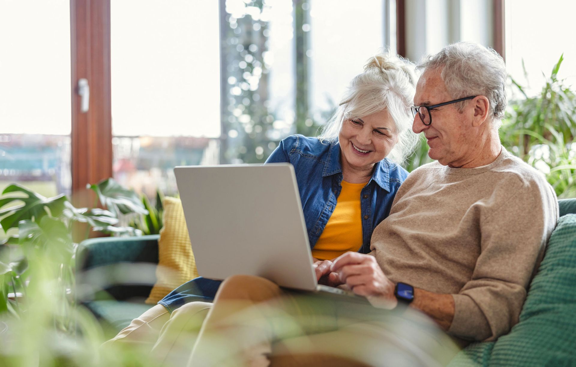 Elderly couple smiling while looking at a laptop together on a couch.