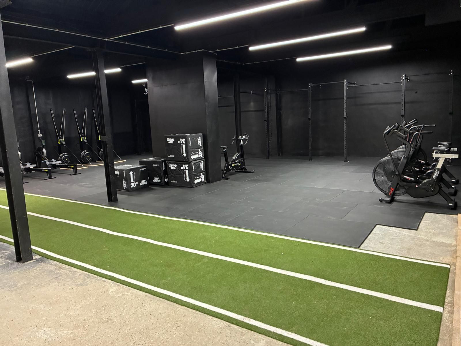 A gym with black walls and ceilings, featuring a green turf track, exercise bikes, and wall-mounted fitness equipment.
