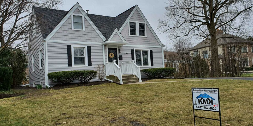 A gray two-story house with a white front door and a promotional yard sign on the lawn during an overcast day.