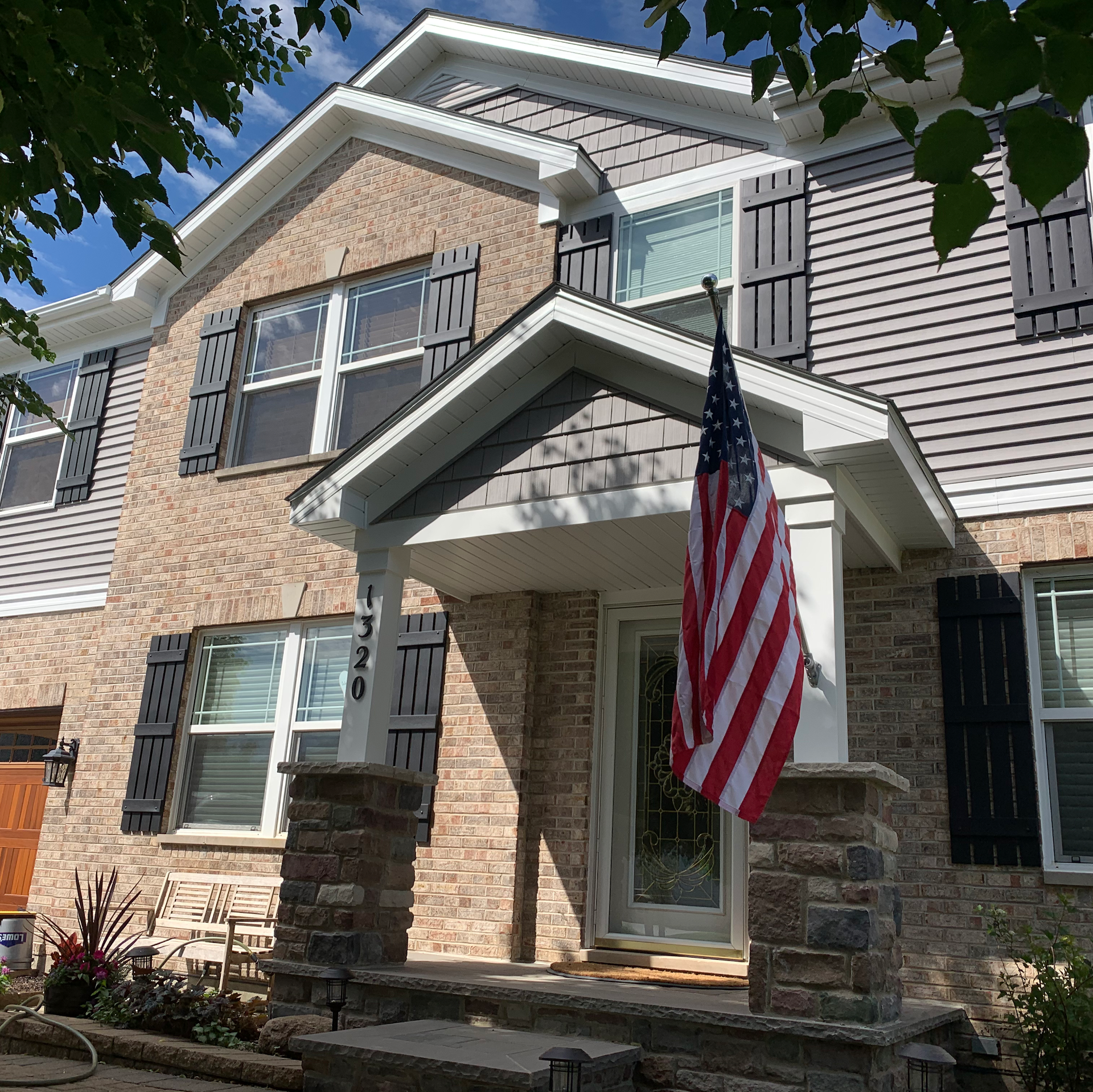 A brick and gray siding two-story house with a front porch, stone columns, dark shutters, and an American flag.