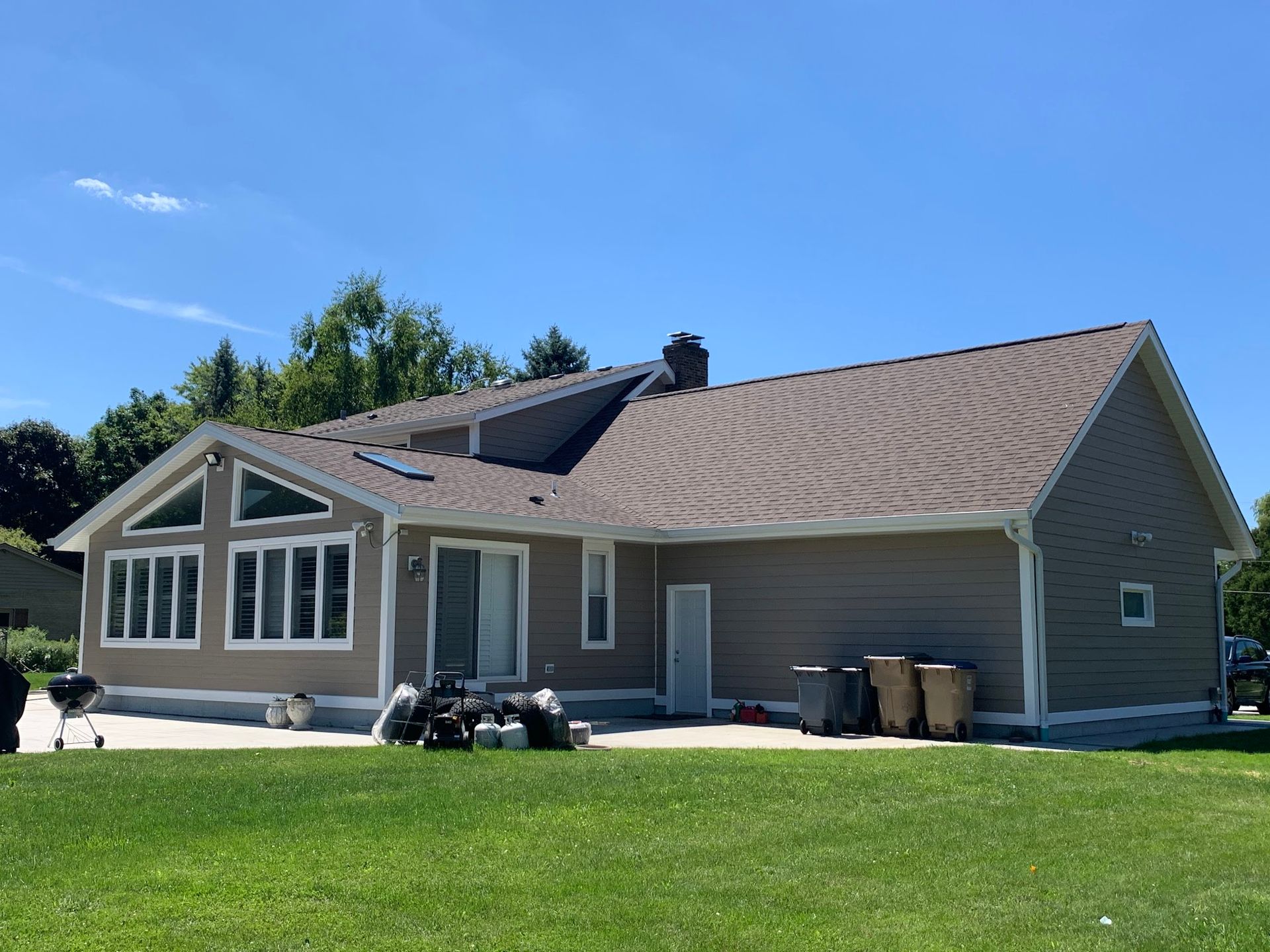 A tan, single-story residential home with brown roof shingles, a sunroom, and a backyard patio under a clear blue sky.