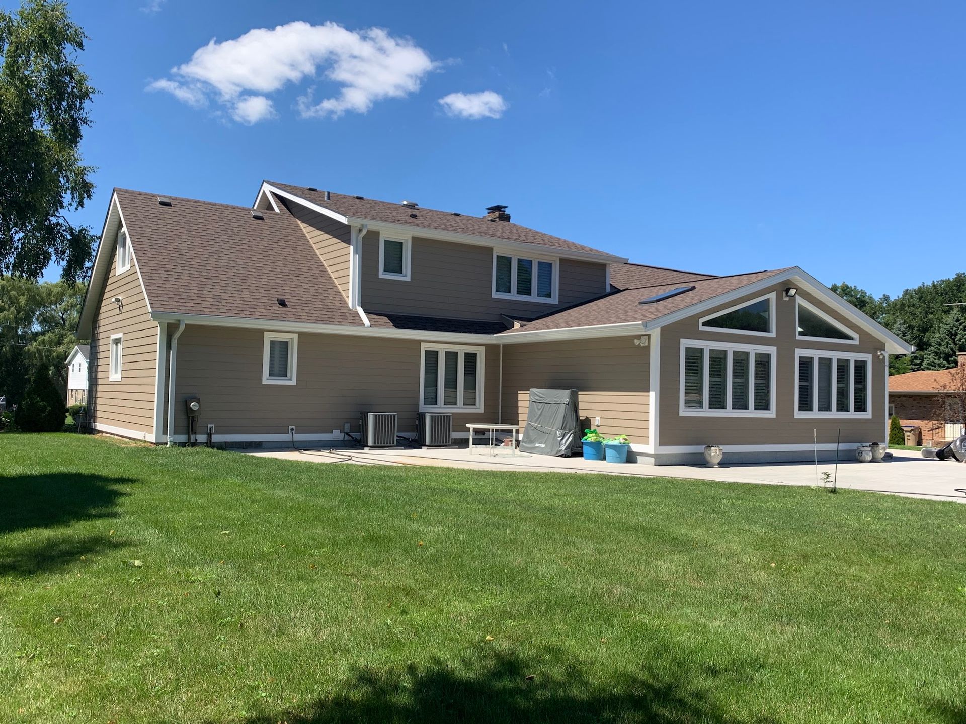 A tan two-story house with a brown shingled roof, large windows, and a paved patio area on a sunny day.