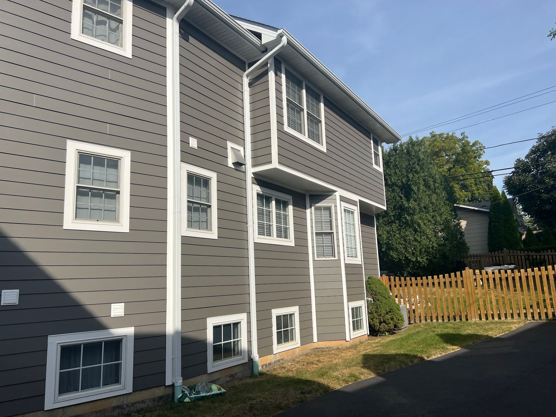 The side view of a gray, multi-story house with white trim, multiple windows, and a wooden fence in the backyard.