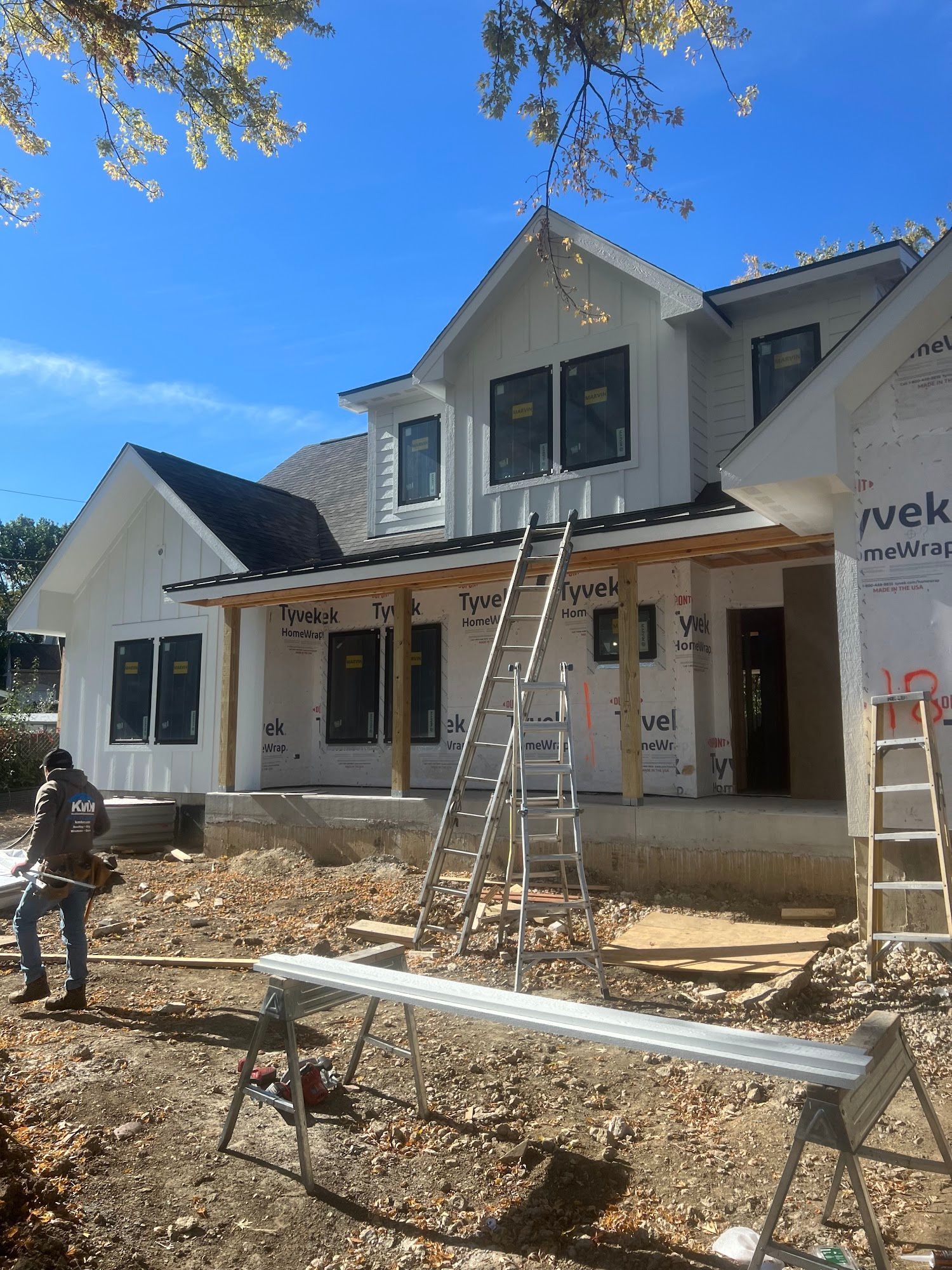 A construction worker stands outside a new, partially finished two-story house with white siding and ladders.