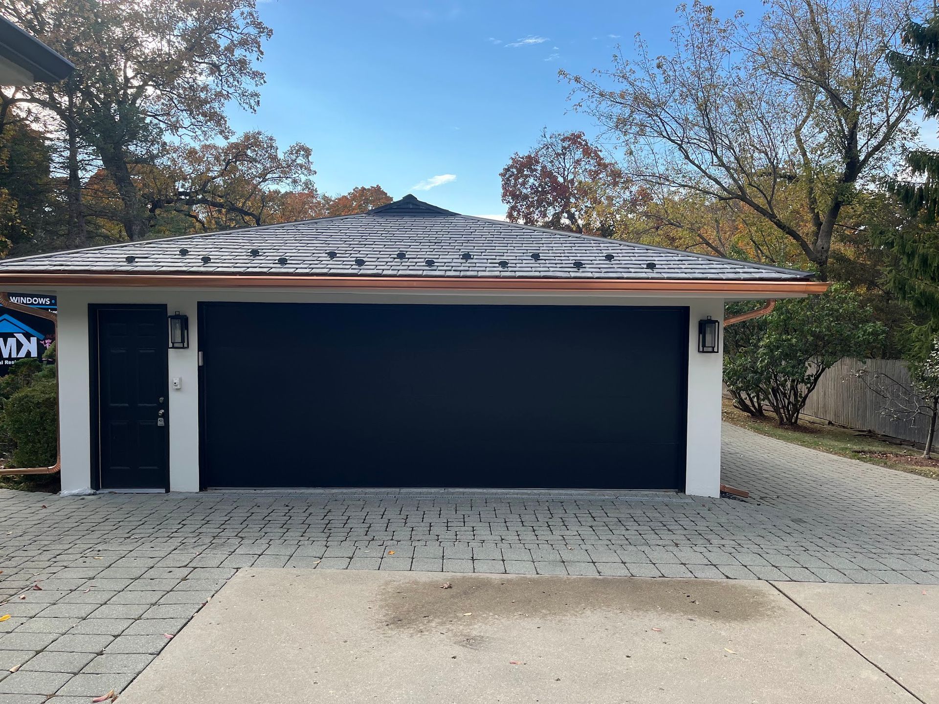 A modern detached garage with a wide black door and matching side entry door, set against a stone paver driveway.