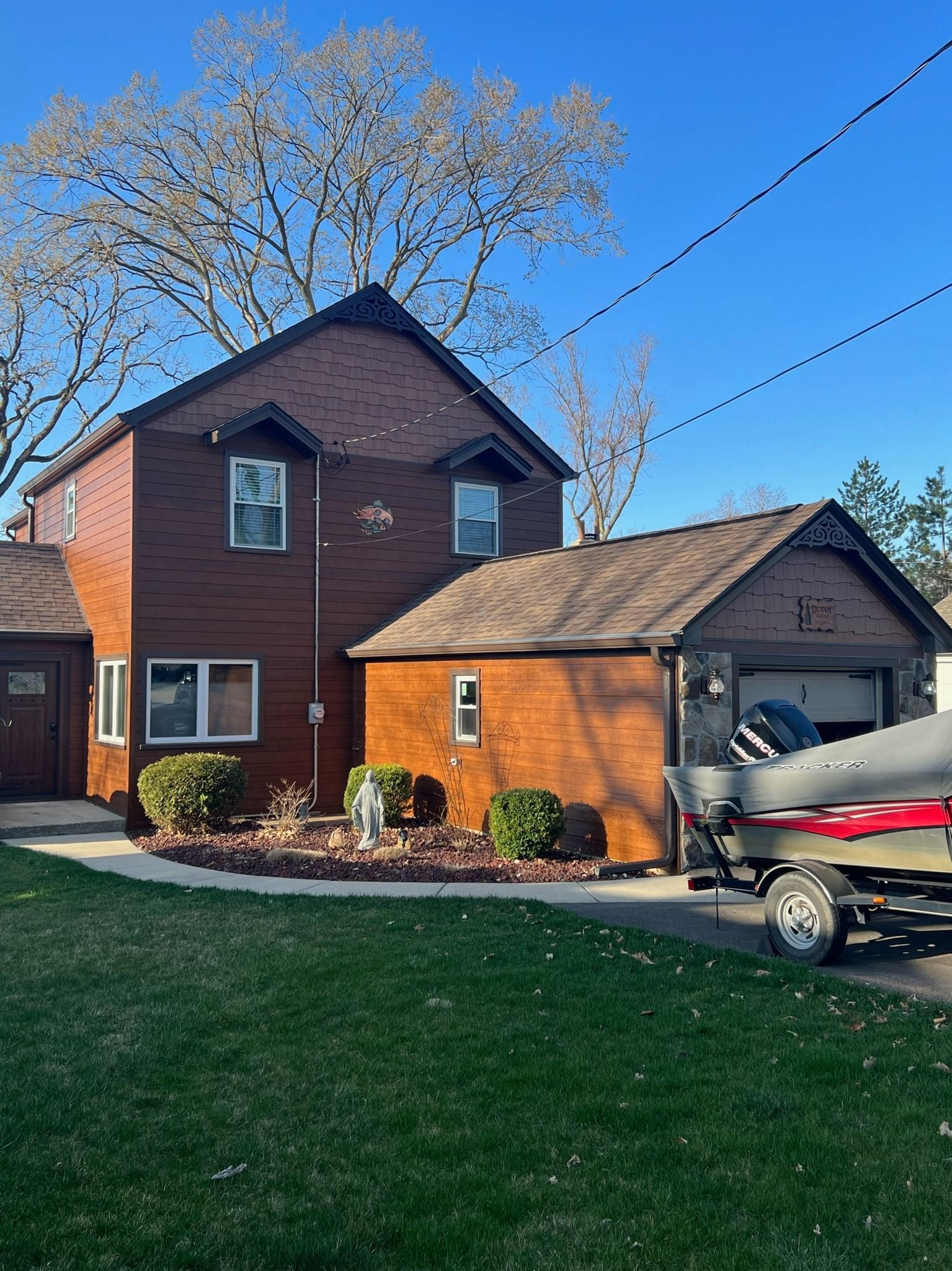 A two-story brown house with a garage and a boat on a trailer parked in the driveway on a sunny day.