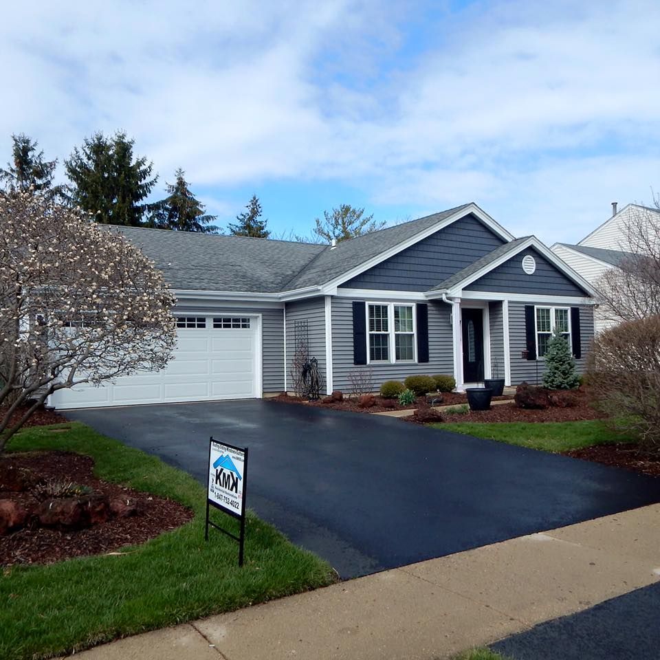 A gray, one-story house with a white garage and trim, featuring a dark front door and a lawn sign in the paved driveway.