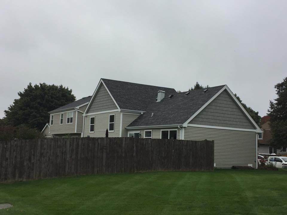 A beige, multi-gabled house with a grey shingled roof, viewed from behind a wooden fence on a grassy lawn under a cloudy sky.
