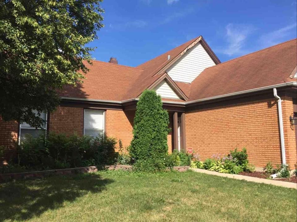 A brick house with a brown shingled roof, a white gabled section, and green landscaping under a bright blue sky.