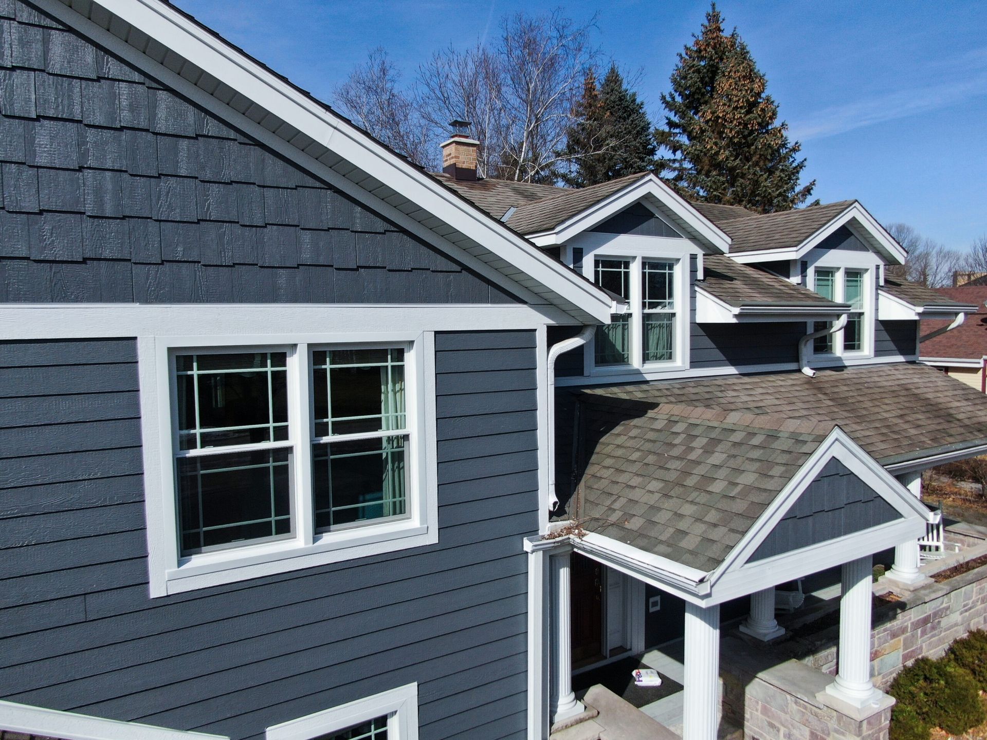 A two-story house with dark grey shingle siding, a stone base, white trim, and multiple gabled roofs under a blue sky.