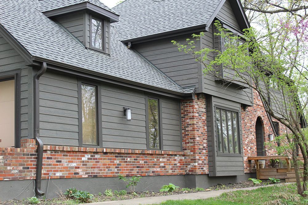 A gray-sided house with a brick base and a multi-gabled roof, partially obscured by a tree, under a clear sky.