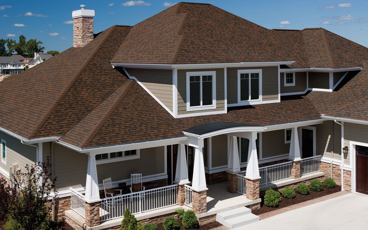 A large, tan suburban house with a brown shingled roof, stone accents, and a white front porch on a sunny day.